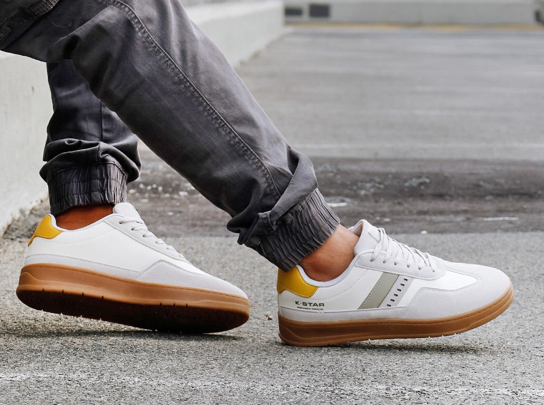 White sneakers with brown soles and yellow accents worn by a person on a concrete surface, with 'K-Star' branding.
