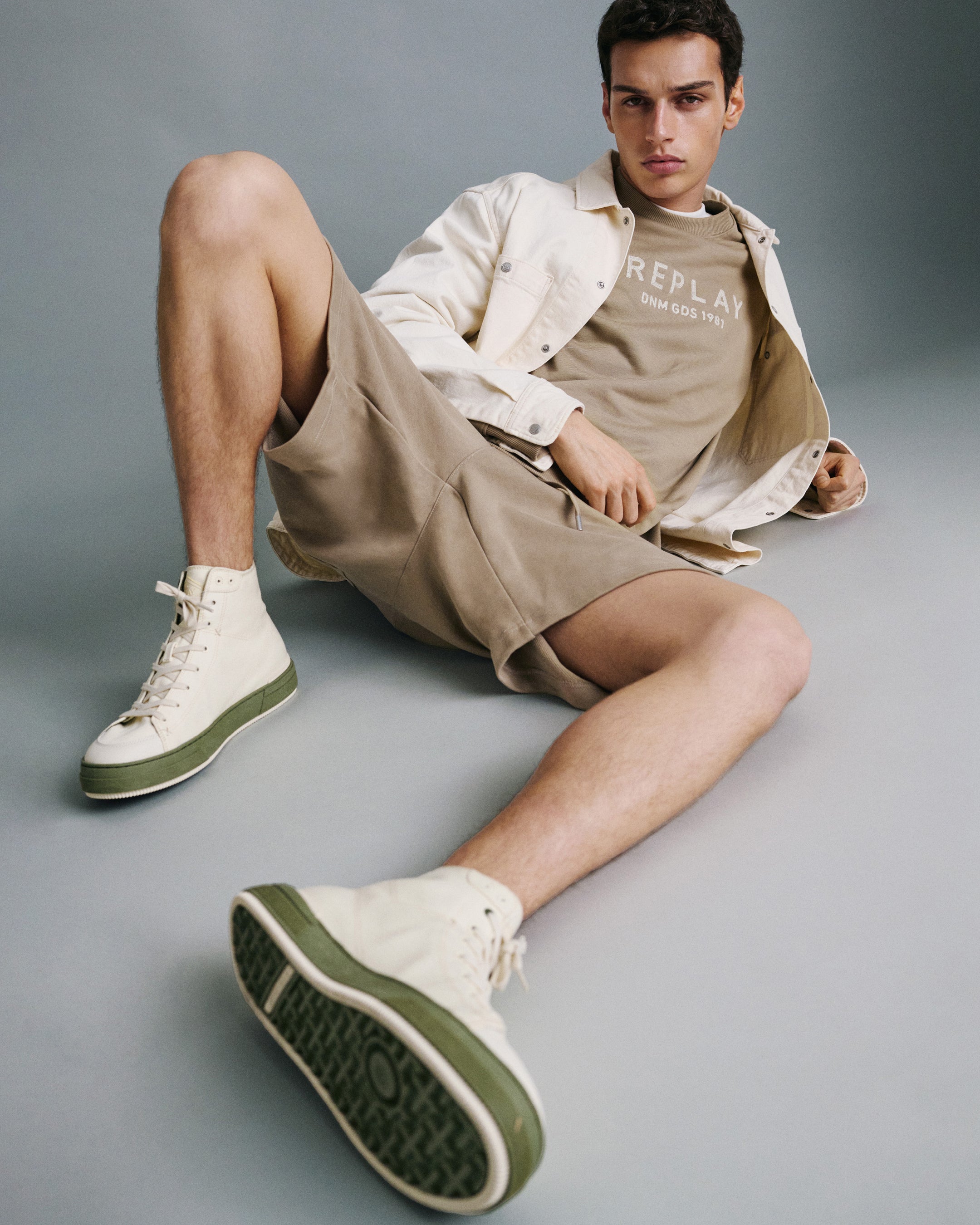 Man sitting on a gray floor wearing beige shorts, white jacket, and green sneakers.